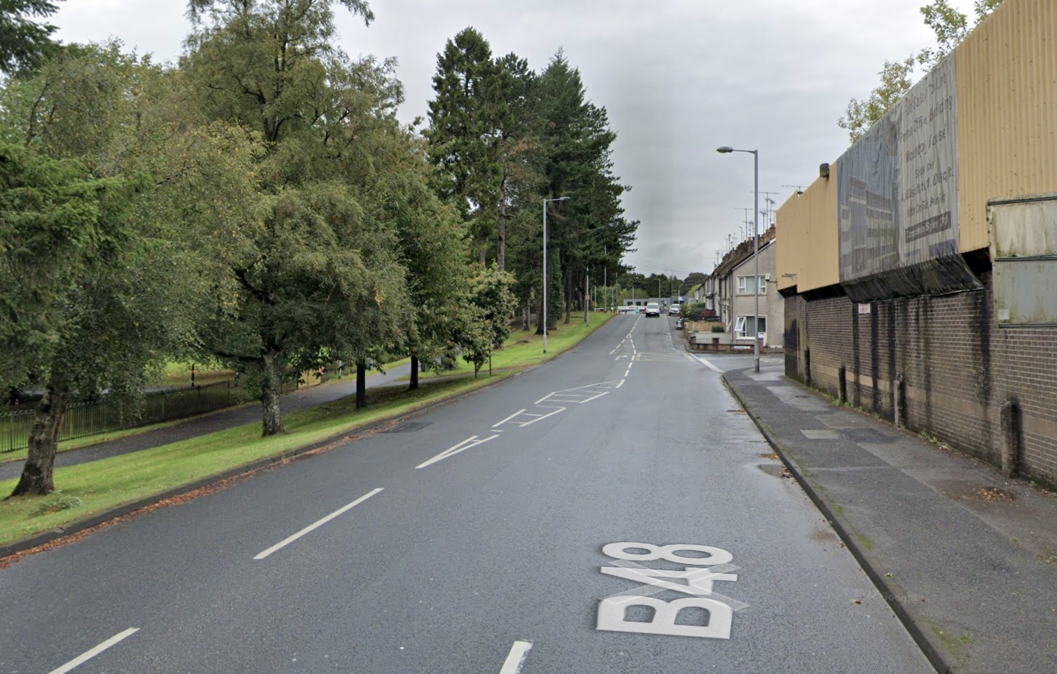 Looking along a two lane road. A high,
                          yellow metal wall of a police station is on
                          the right. On the left is a park with a line
                          of trees beside the road, and footpath running
                          parallel to the road behind the trees.