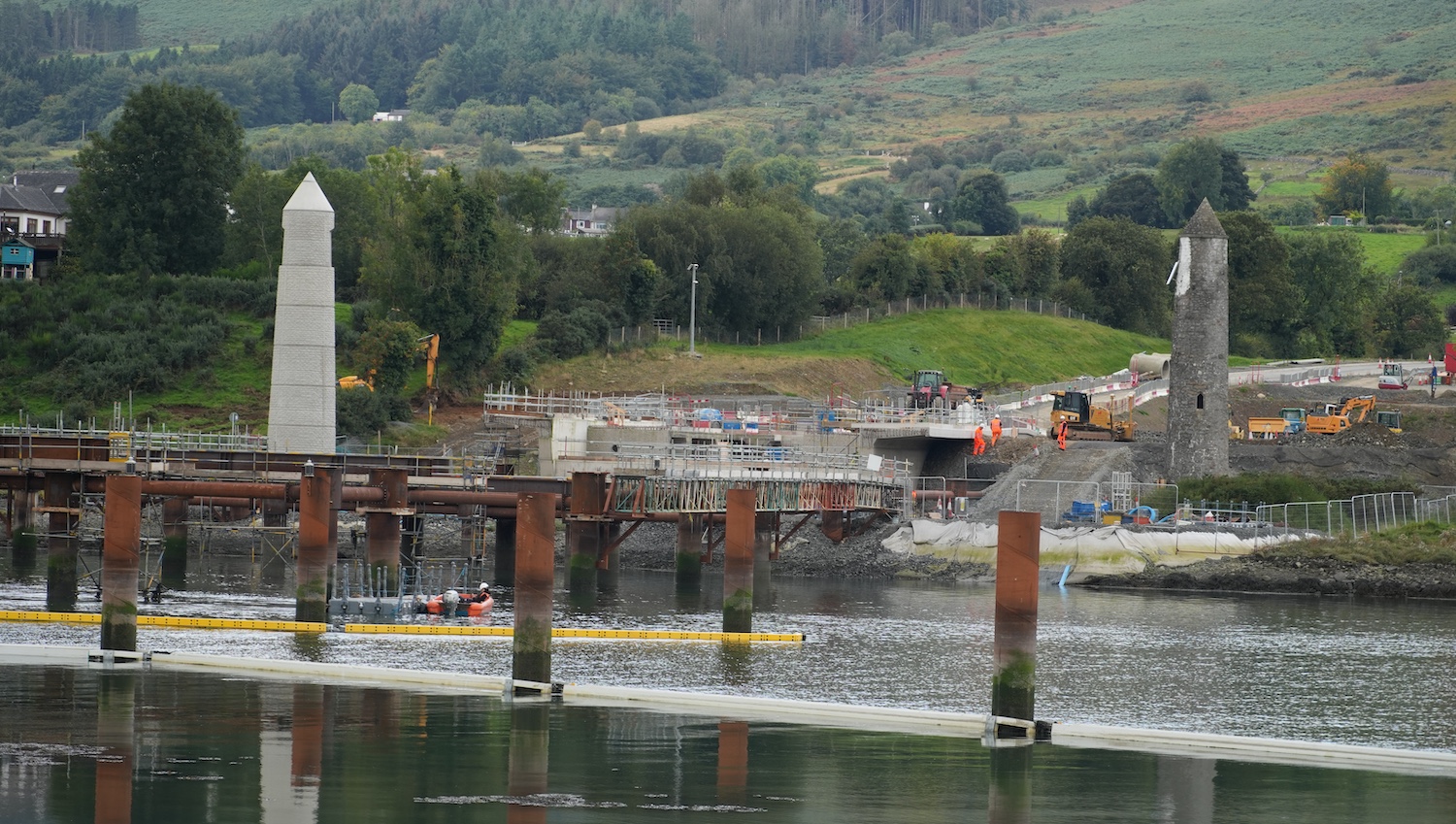 View across the river of a concrete
abutment stopping suddently and then a gap
where the metal bridge section will go. There
are metal posts and frames in the river in the
foreground.